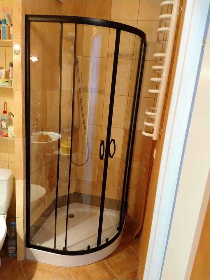 Narożna kabina prysznicowa z czarną ramą i przezroczystymi szybami w łazience z beżowymi płytkami, widoczna toaleta i półka z kosmetykami.