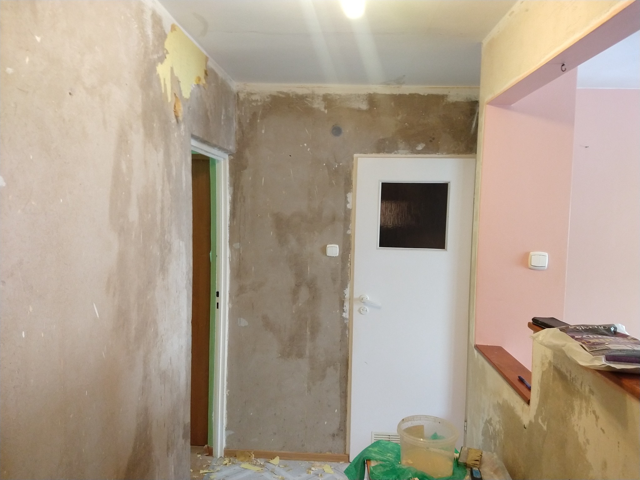 Przygotowanie ścian do prac wykończeniowych: usunięte stare powłoki, ściany częściowo zagruntowane, widoczne resztki tapet i farb, plastikowe wiadro i pędzel na podłodze, białe drzwi z małym...