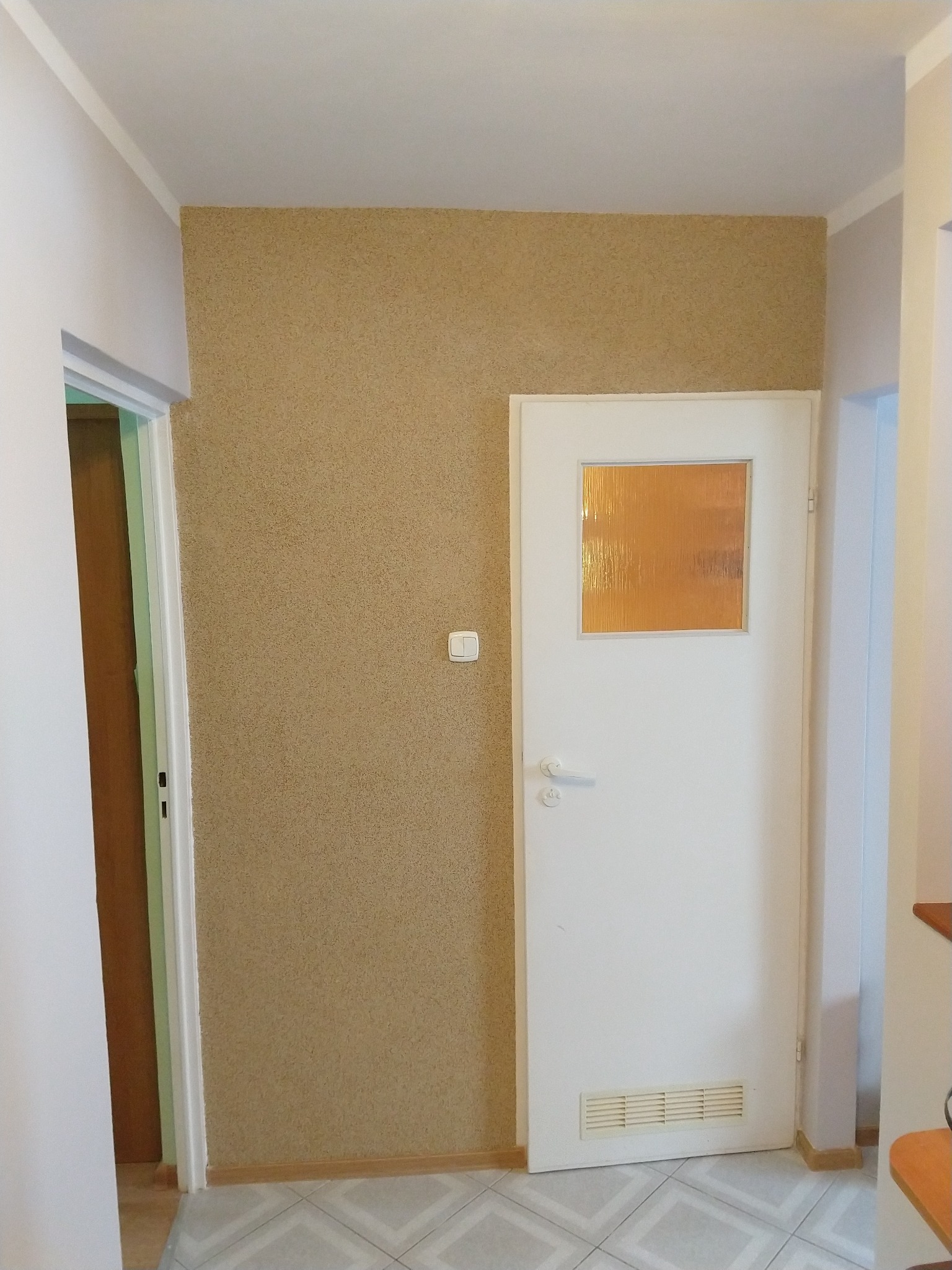 Korytarz po odświeżeniu: ściana pokryta beżową tapetą strukturalną, białe drzwi z szybką z pomarańczowego szkła, jasne płytki podłogowe w geometryczny wzór.