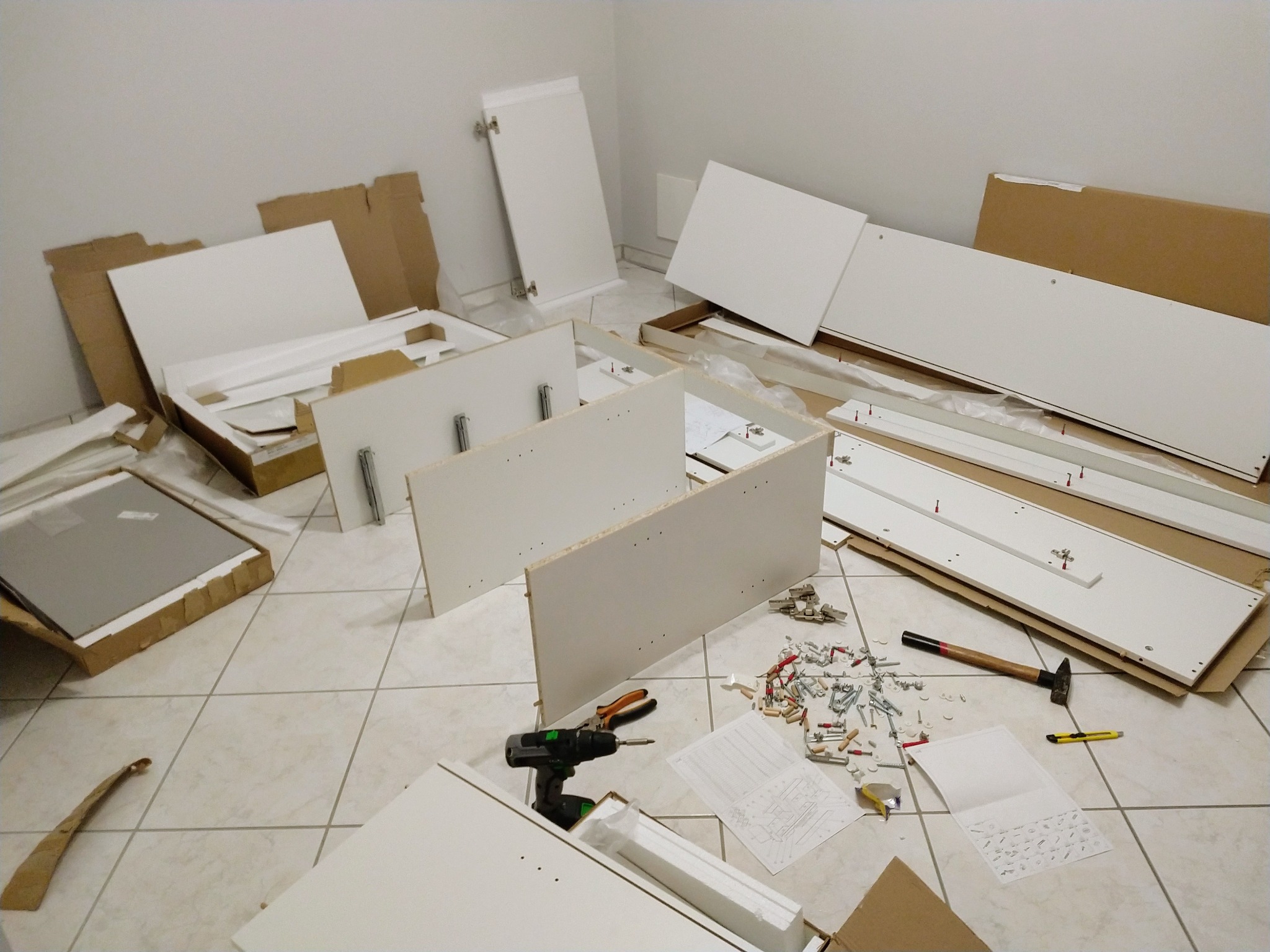 Rozpakowane elementy białej szafki z instrukcją montażu, wkrętakiem, młotkiem i drobnymi elementami montażowymi rozłożone na kafelkach podłogowych, przygotowane do składania.