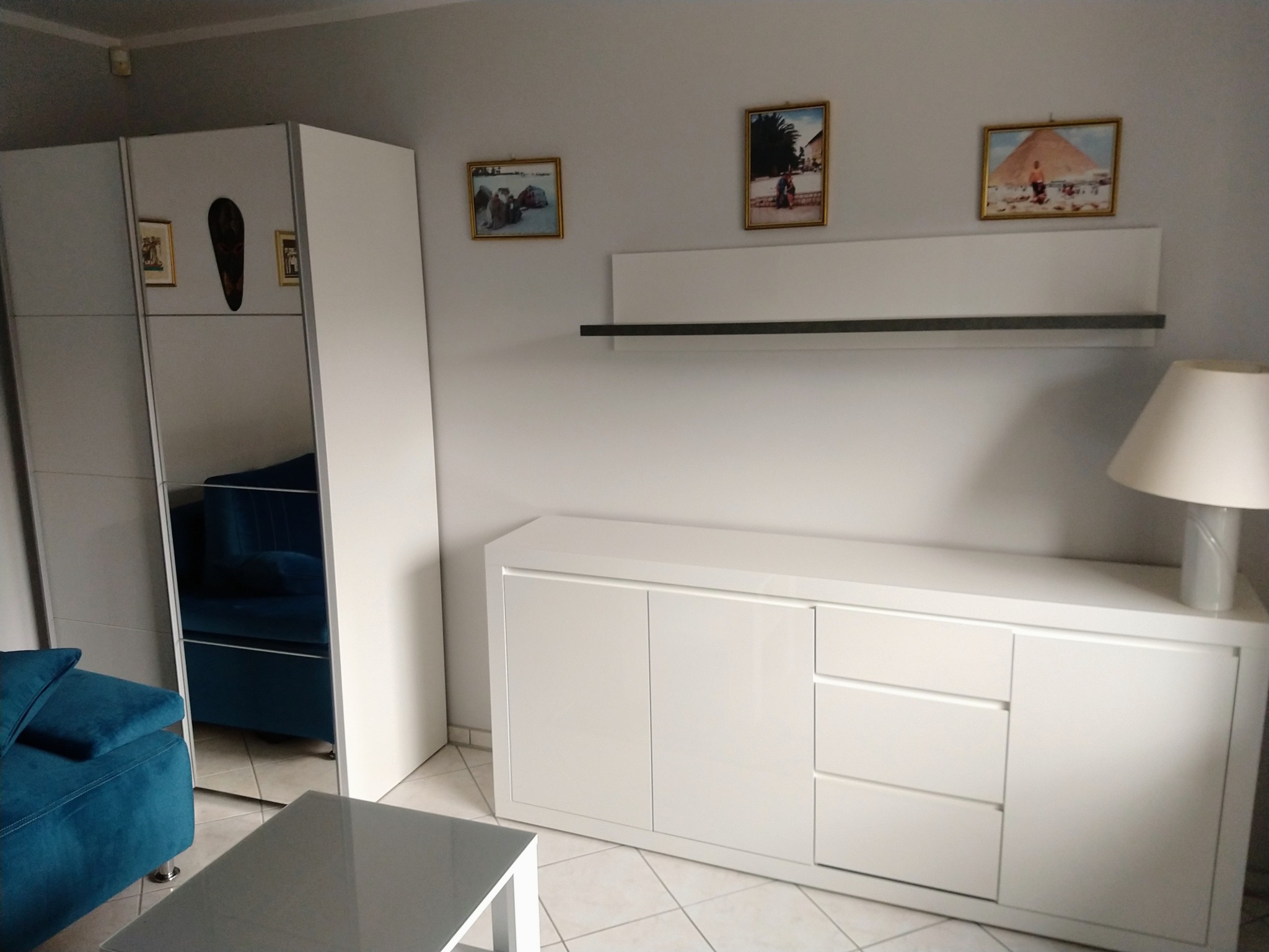 Jasne wnętrze pokoju dziennego z białymi meblami: szafa z lustrem odbijająca turkusową sofę, komoda pod telewizor, szara ława i dekoracje ścienne w postaci ramek z obrazami.