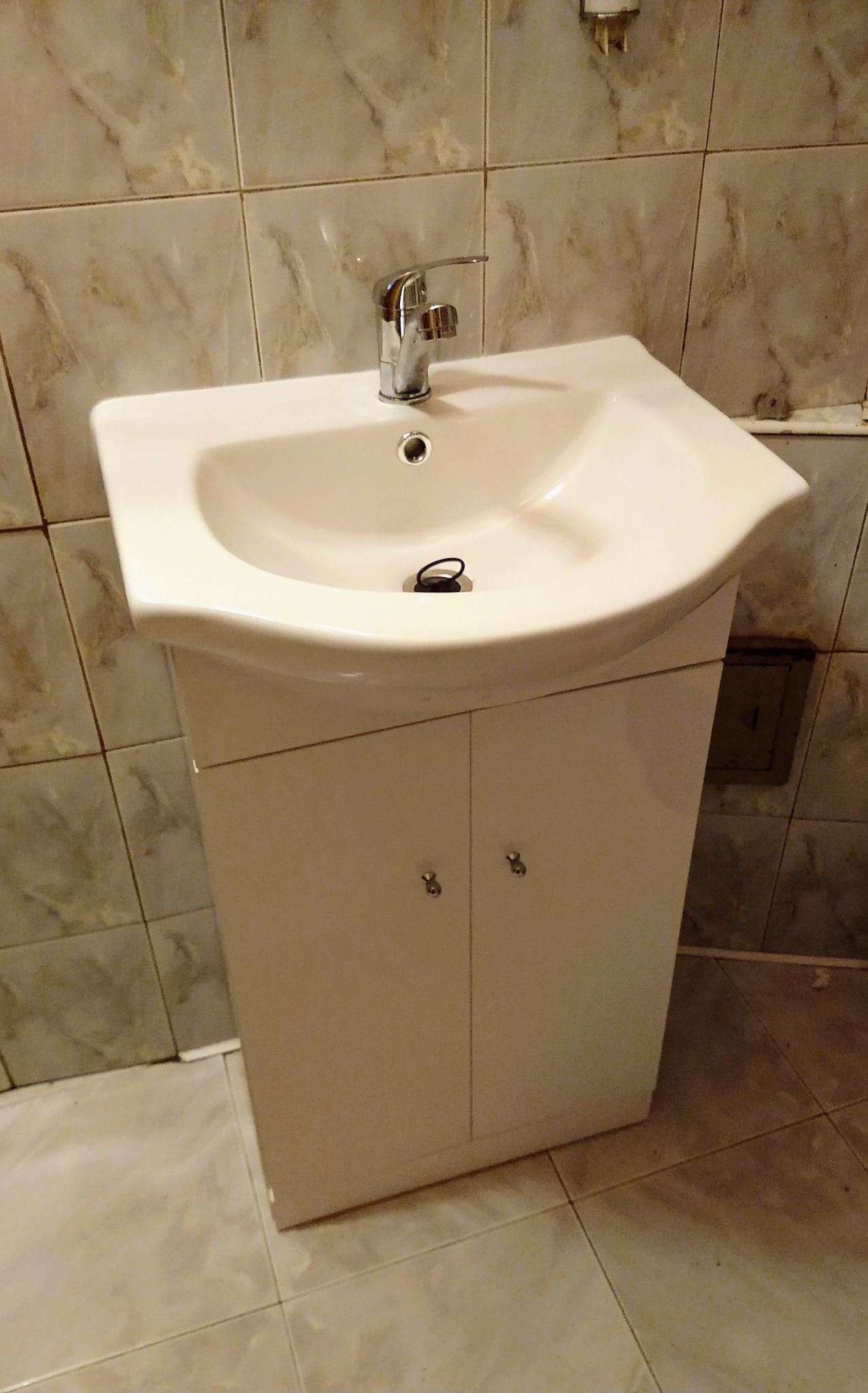 Biała umywalka z szafką w łazience z marmurkowymi płytkami, widoczny chromowany kran i czarny korek spustowy, minimalizm.