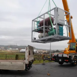 Żółty dźwig samochodowy opuszcza w stalowej klatce urządzenie przemysłowe składające się z czerwonego zbiornika, zielonego silnika i białej sprężarki na przyczepę ciężarową na tle zabudowań...