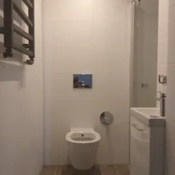 Nowoczesna łazienka z podwieszaną toaletą, białą armaturą, grzejnikiem drabinkowym i beżowymi płytkami podłogowymi.