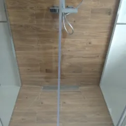 Kabina prysznicowa z drewnopodobnymi płytkami, widoczny odpływ liniowy w podłodze i bateria natryskowa.