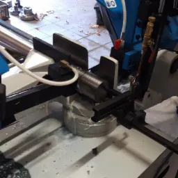 Proces cięcia metalu na pile taśmowej z użyciem chłodziwa, widoczne wióry na podłodze warsztatu.