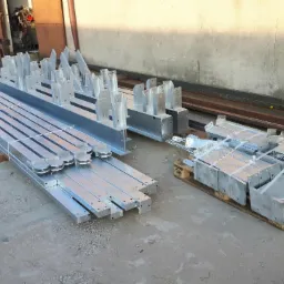 Elementy konstrukcyjne ze stali ocynkowanej, przygotowane do montażu, ułożone w wiązki i na palecie, widoczne otwory montażowe i mocowania.