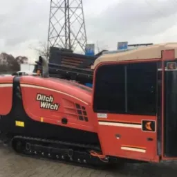 Pomarańczowo-czarna maszyna do przewiertów sterowanych marki Ditch Witch JT30 na gąsienicach, widoczna z boku na tle betonowej nawierzchni i słupa wysokiego napięcia.