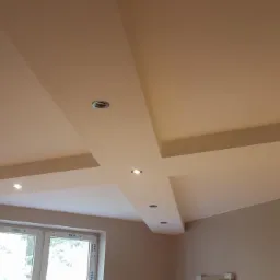 Wykończony sufit podwieszany z płyt kartonowo-gipsowych, z wbudowanymi punktami świetlnymi, pomalowany na jednolity jasny kolor, widok z dołu na okno.