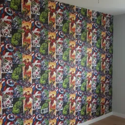 Ściana pokoju dziecięcego pokryta tapetą z motywami komiksowymi Marvela: Iron Man, Hulk, Kapitan Ameryka. Obok ściana pomalowana na szaro i okno z białymi ramami.