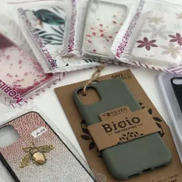 Różnorodne etui na telefony, w tym silikonowe, brokatowe i z motywami florystycznymi, prezentowane na białym tle. Jedno z etui, wykonane z materiałów roślinnych, owinięte papierową opaską z logo...