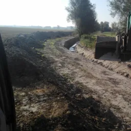 Koparka pogłębia rów melioracyjny z wodą, widok z kabiny operatora na wykopany kanał w krajobrazie rolniczym.