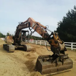 Żółta koparka gąsienicowa z operatorem w kabinie, na piaszczystym terenie, obok białego płotu i drzew.