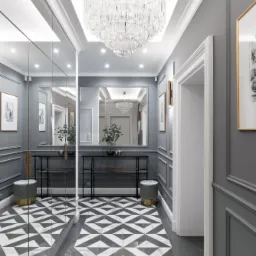 Elegancki przedpokój w odcieniach szarości z geometryczną podłogą, lustrzaną szafą i kryształowym żyrandolem. W tle widoczne obrazy w złotych ramach i dekoracyjne sztukaterie.
