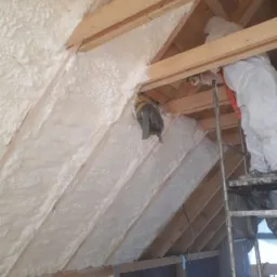 Aplikacja białej piany izolacyjnej na skośnym poddaszu, widoczne drewniane krokwie, pracownik w kombinezonie ochronnym na drabinie, częściowo zakryta folią budowlaną