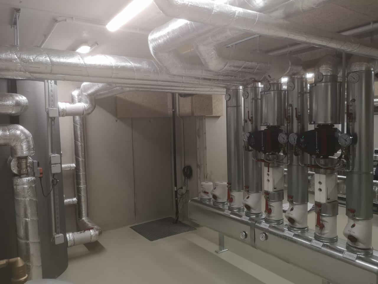 Izolowane rury instalacji grzewczej i hydraulicznej w piwnicy, widoczne pompy obiegowe i kolektor rozdzielczy, szare ściany i podłoga, oświetlenie sufitowe.