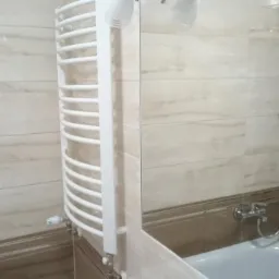 Wykończona łazienka z beżowymi płytkami imitującymi marmur, grzejnikiem drabinkowym i kabiną prysznicową z widocznym zestawem natryskowym.