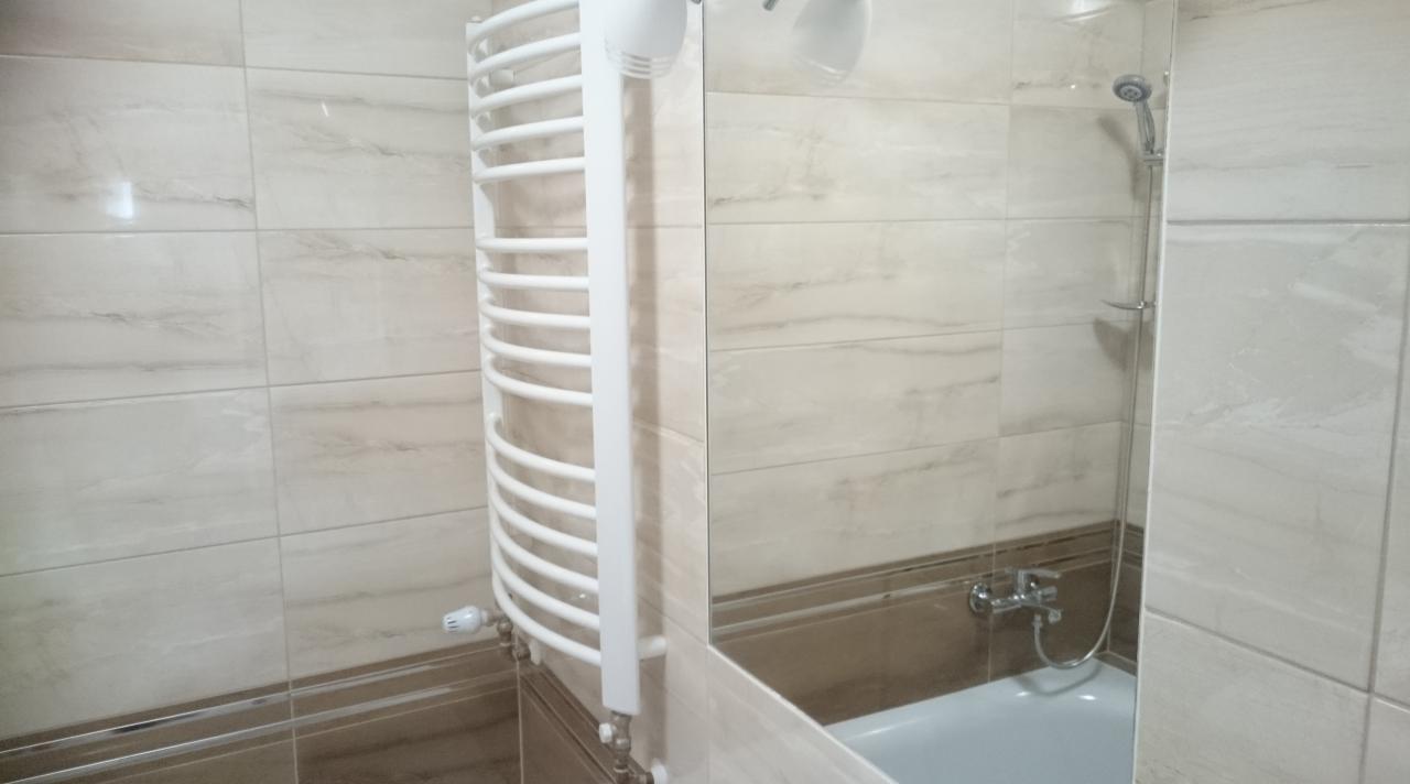 Wykończona łazienka z beżowymi płytkami imitującymi marmur, grzejnikiem drabinkowym i kabiną prysznicową z widocznym zestawem natryskowym.