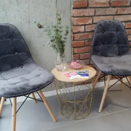 Poczekalnia: Dwa szare, welurowe krzesła z drewnianymi nogami, stolik kawowy z metalowym koszem, wazon z gałązkami i ulotki informacyjne na tle ceglanej ściany.