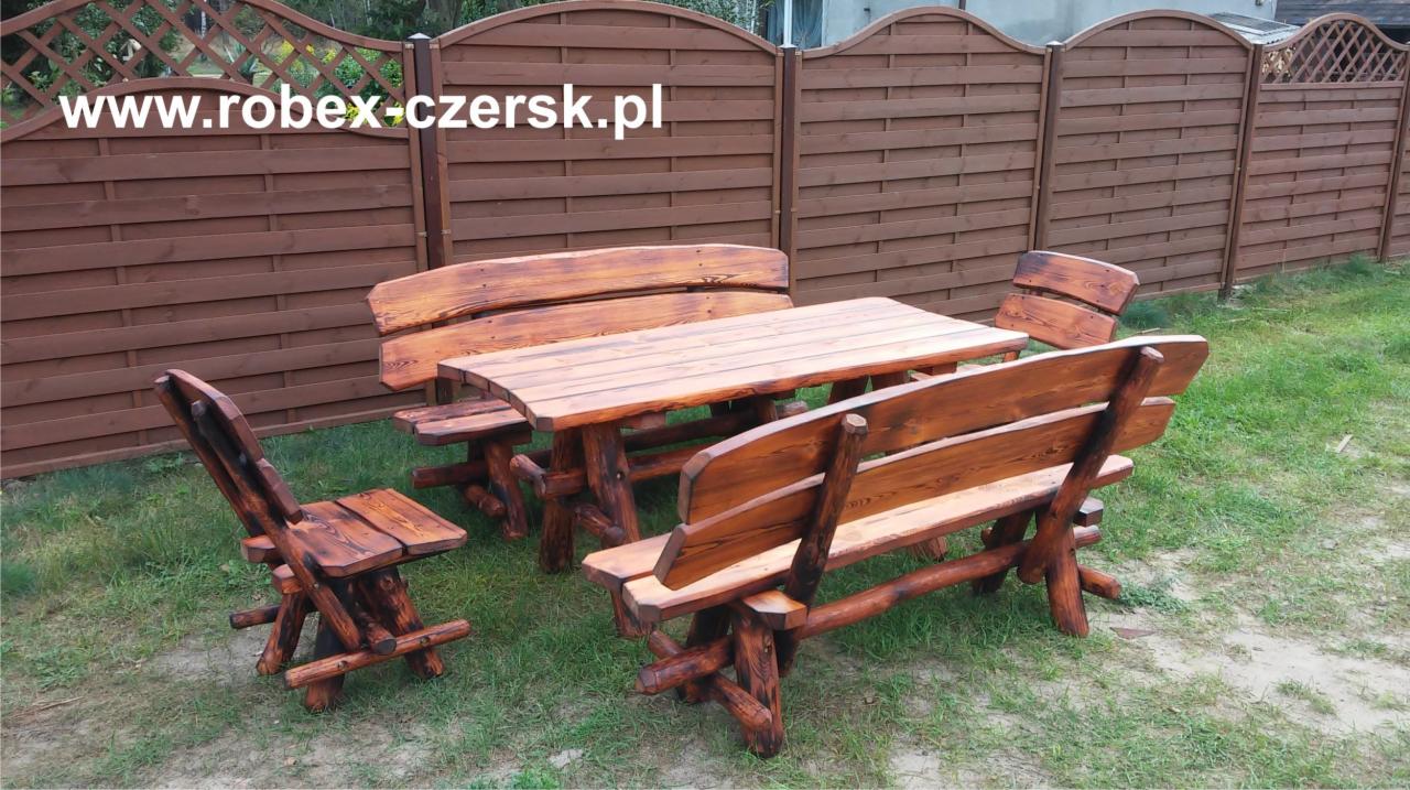 Rustykalny zestaw mebli ogrodowych z litego drewna: stół z ławkami i krzesłami, na tle brązowego płotu lamelowego i zielonego trawnika.