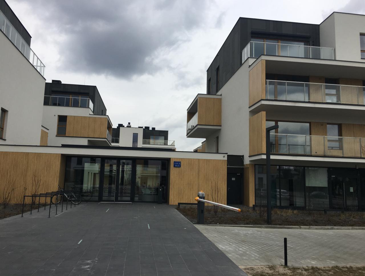 Nowoczesny budynek apartamentowy z drewnianymi akcentami elewacji i szklanymi balkonami, widok z poziomu chodnika, pochmurne niebo w tle.