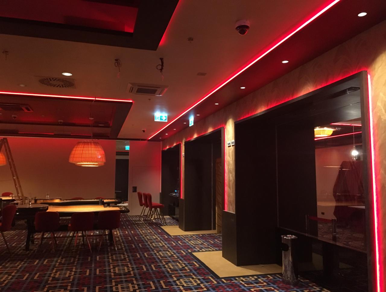 Nowoczesne wnętrze z czerwonym oświetleniem LED, wzorzystą wykładziną i ciemnymi elementami wykończenia, widoczne stoliki i krzesła w tle.