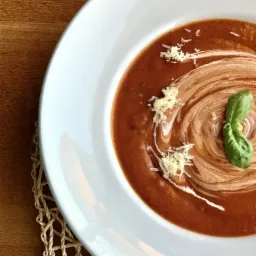 Krem pomidorowy w białym talerzu, udekorowany kleksem śmietany, listkiem bazylii i posypany startym serem, na drewnianym stole z plecioną podkładką.