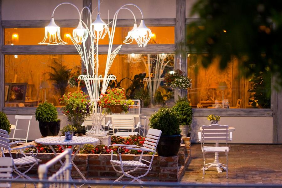 Ustawione na zewnątrz białe meble ogrodowe: stoliki z kwiatami, krzesła i dekoracyjna lampa, na tle okna budynku z oświetlonym wnętrzem.