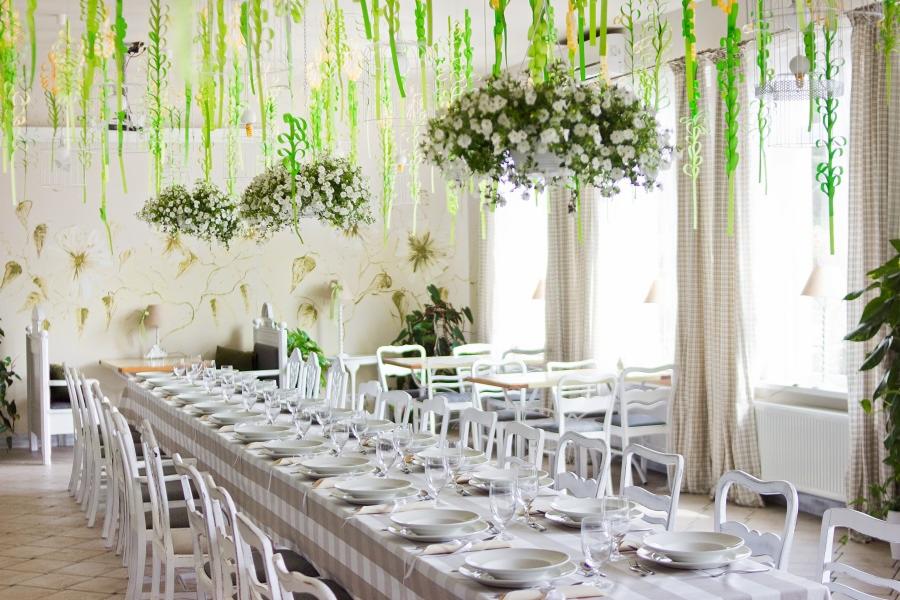 Uroczysta sala bankietowa z długim stołem nakrytym zastawą, dekoracjami kwiatowymi pod sufitem i zielonymi wstążkami, jasne wnętrze.