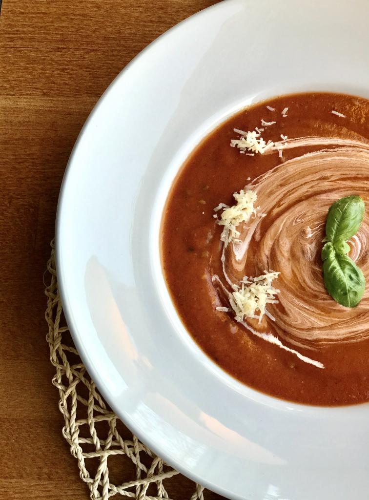 Krem pomidorowy w białym talerzu, udekorowany kleksem śmietany, listkiem bazylii i posypany startym serem, na drewnianym stole z plecioną podkładką.