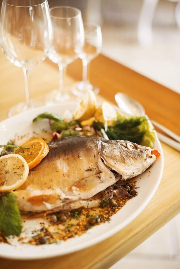 Cała pieczona ryba na białym talerzu, udekorowana cytrusami i ziołami, z trzema kieliszkami w tle.