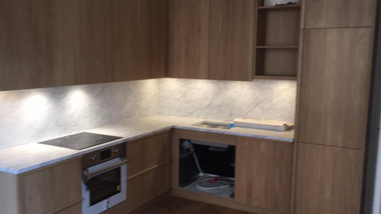 Fragment kuchni z drewnianymi szafkami, marmurowym blatem i oświetleniem podszafkowym, piekarnik w zabudowie, brakujące drzwiczki szafki pod zlewem.