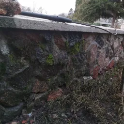 Kamienny murek z betonowym daszkiem, częściowo porośnięty mchem, z widocznym systemem nawadniania w postaci czarnej rury z otworami, leżącej na daszku.