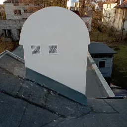 Odnowiony komin z dwoma kratkami wentylacyjnymi na dachu pokrytym papą, widok z góry na tle zabudowy mieszkalnej i drzew bez liści.