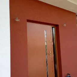 Brązowa elewacja z tynkiem strukturalnym, drewniane drzwi wejściowe z przeszkleniem, zwinięty przedłużacz leżący na drewnianym podeście.