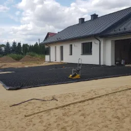 Przygotowanie podłoża pod taras: rozłożona czarna siatka na piasku, ubijarka wibracyjna, taczka i kupki piachu w tle, dom w stylu nowoczesnym z garażem w trakcie wykończenia.