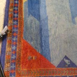 CleanIT - Czyszczenie kolorowego dywanu wzorzystego, widoczna końcówka odkurzacza piorącego, jasne drewniane panele podłogowe, widoczny fragment czarnego plecaka w tle.