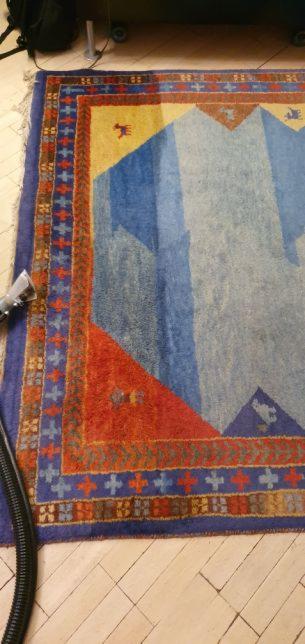 Czyszczenie kolorowego dywanu wzorzystego, widoczna końcówka odkurzacza piorącego, jasne drewniane panele podłogowe, widoczny fragment czarnego plecaka w tle.