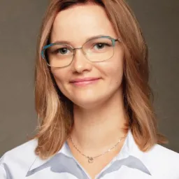 Portret uśmiechniętej kobiety w okularach z kolorowymi oprawkami i białej koszuli, z delikatnym naszyjnikiem w kształcie serca na tle szarego studia.