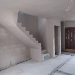 Wnętrze domu w trakcie prac wykończeniowych: świeżo nałożone tynki na ścianach i schodach, widoczne przewody instalacyjne na podłodze, surowa klatka schodowa z widocznymi stopniami.