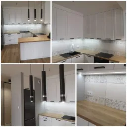 Nowoczesna, jasna kuchnia z białymi szafkami, drewnianym blatem i czarnymi, cylindrycznymi lampami wiszącymi. Dekoracyjne płytki nad blatem dodają charakteru.