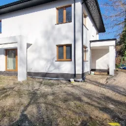 Biały dom z brązowymi oknami w słoneczny dzień. Widoczna świeżo pomalowana elewacja i czarne rynny. Na ziemi widać resztki liści i trawy.