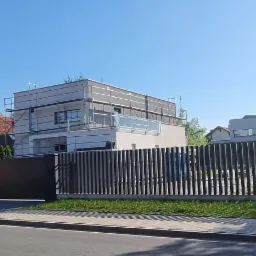 Dom w trakcie ocieplania elewacji, widoczne rusztowania. Nowoczesna architektura, jasna kolorystyka budynku. Na pierwszym planie ogrodzenie i fragment ulicy.