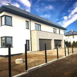 Krishome - Nowoczesny dom jednorodzinny z białymi ścianami, szarymi oknami PCV i segmentową bramą garażową w trakcie montażu ogrodzenia z czarnych słupków na tle błękitnego nieba.