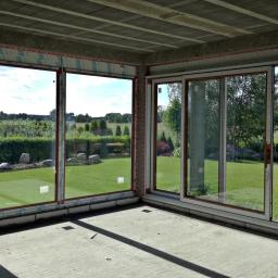 Krishome - Surowy stan wnętrza z dużymi oknami PCV, przez które widać zadbany ogród z trawnikiem, krzewami i drzewami; widoczne profile okienne z naklejkami ochronnymi.