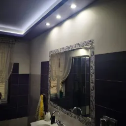 Nowoczesna łazienka z podwieszanym sufitem z oświetleniem LED, ciemnymi płytkami i dużym lustrem z dekoracyjną ramą.