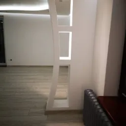 Biała, dekoracyjna ścianka działowa z podświetlanymi wnękami, dzieląca przestrzeń w nowocześnie wykończonym wnętrzu z drewnopodobną podłogą i grzejnikiem żeliwnym.