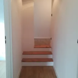 Nowoczesne wnętrze z drewnianą podłogą, stopniami schodów z litego drewna i białymi ścianami oświetlonymi punktowo, widok z perspektywy wejścia do pomieszczenia.