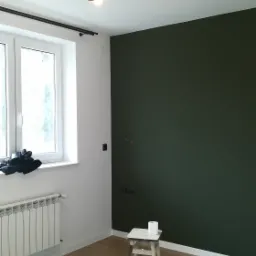 Pokój po malowaniu jednej ściany na ciemnozielony kolor, widoczny stołek z rolką papieru toaletowego, lampa sufitowa z trzema reflektorami, okno z grzejnikiem, podłoga z paneli oraz buty i torba...