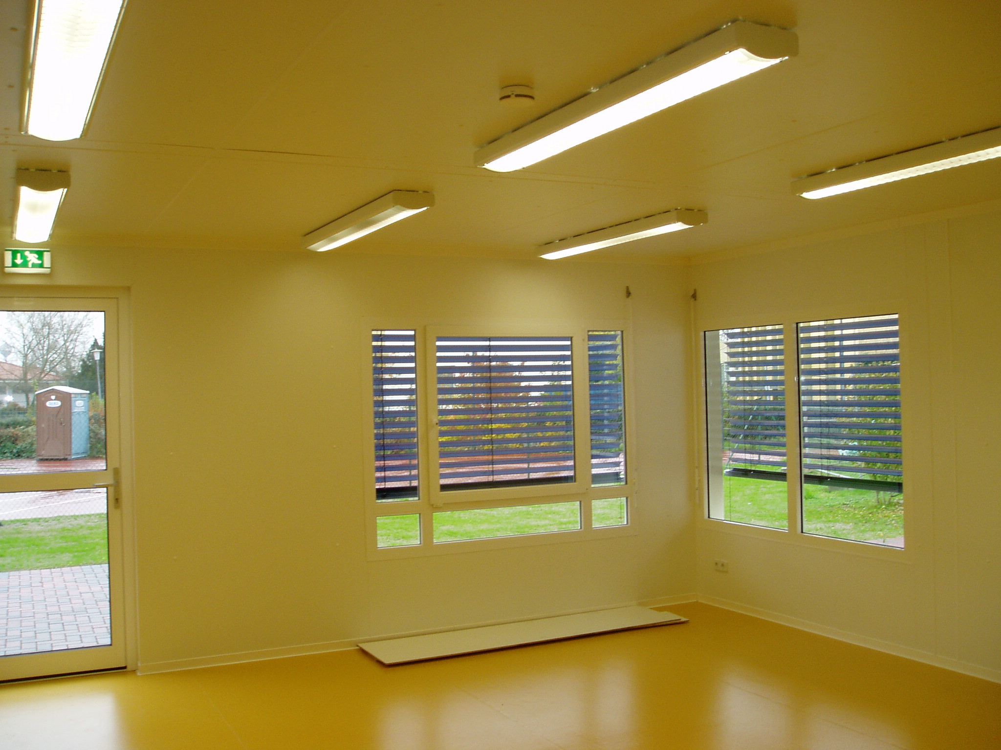 Wnętrze modułowego budynku z żółtą podłogą, oświetleniem jarzeniowym i oknami z żaluzjami, widok na zewnątrz z toaletą przenośną.